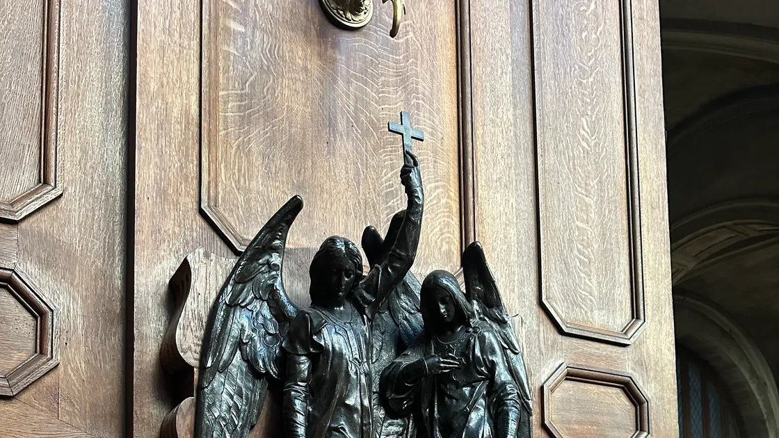 Pia de água benta de bronze com dois anjos segurando uma cruz, sobre painel de madeira entalhada numa igreja
