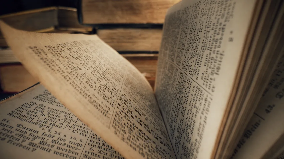 Macro de páginas antigas de uma Bíblia aberta, tipografia gótica, com outros livros de capa dura desfocados ao fundo