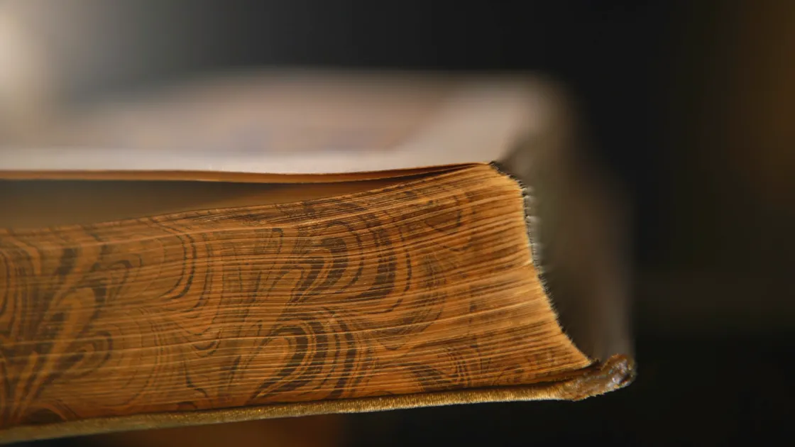Detalhe macro da borda dourada e marmorizada de um livro antigo de capa dura, sobre fundo escuro desfocado