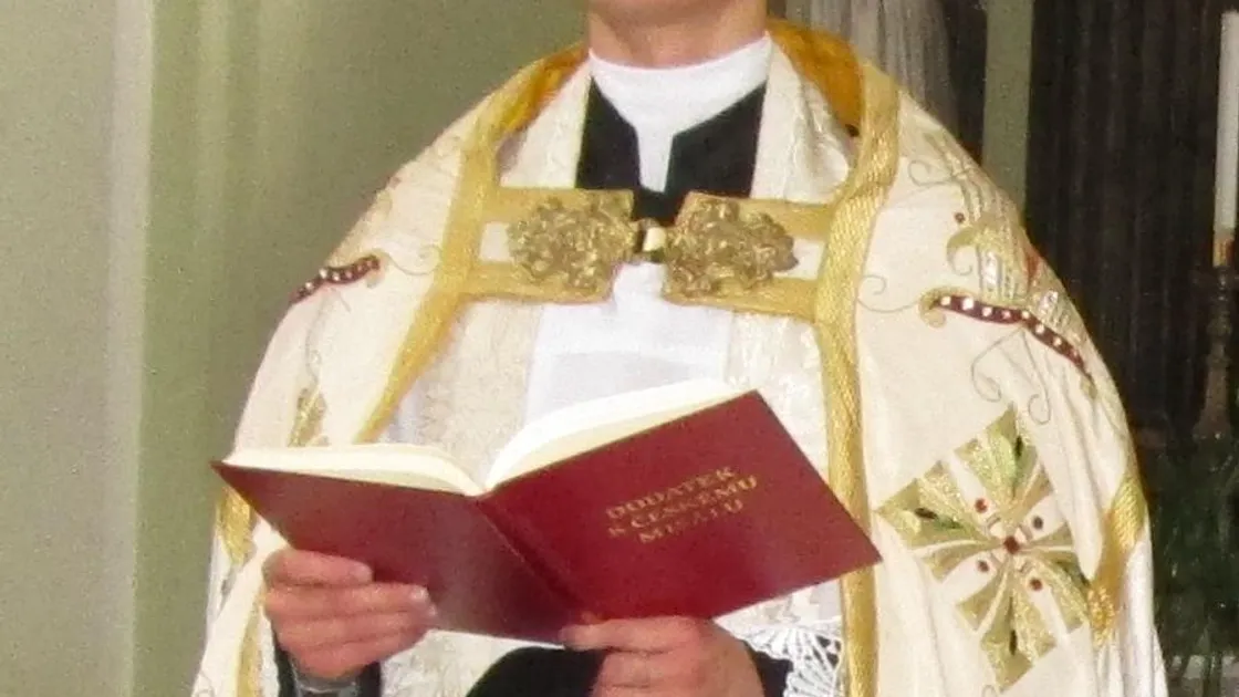 Padre paramentado com capa branca bordada em dourado, segurando um missal aberto durante celebração