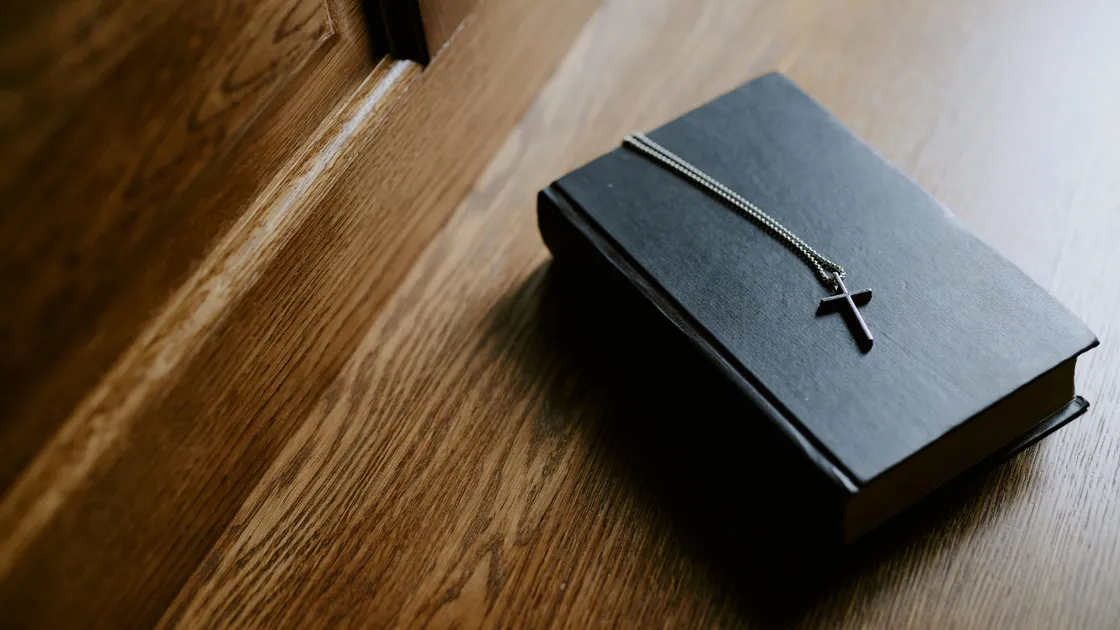 Livro de capa preta fechado sobre piso de madeira, com um colar de crucifixo prateado apoiado por cima