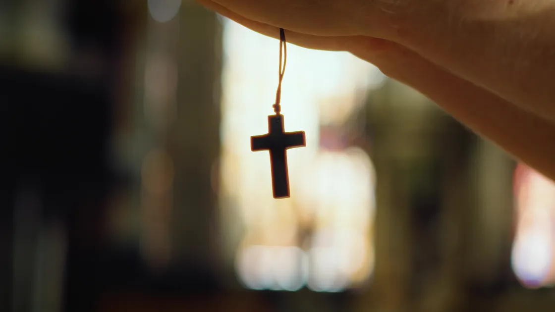 Crucifixo de terço pendurado entre os dedos de uma mão, em contraluz dentro de uma igreja