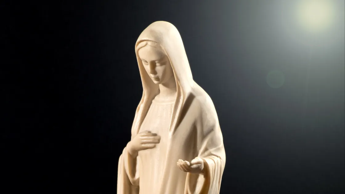 Imagem devocional de Nossa Senhora em resina clara, mãos abertas, iluminada por um facho de luz sobre fundo escuro