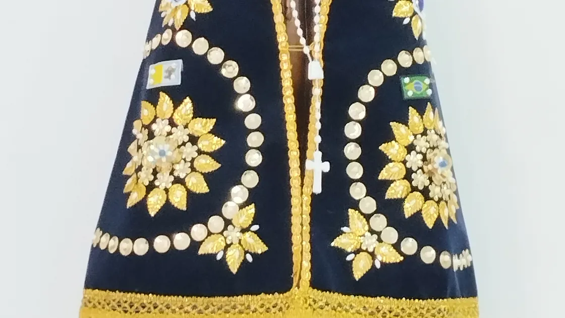 Imagem de Nossa Senhora Aparecida com manto azul bordado a dourado, rosto em terracota escura, sobre fundo branco.