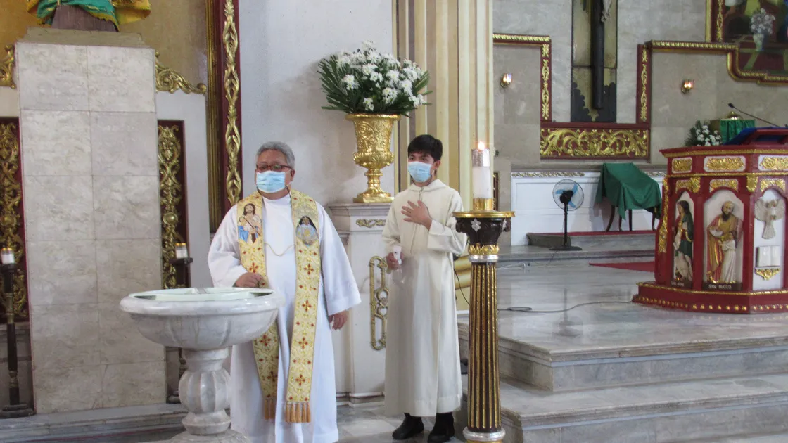 Padre ao lado de uma pia batismal de mármore e um coroinha segurando um círio aceso, dentro de uma igreja católica decorada em dourado.