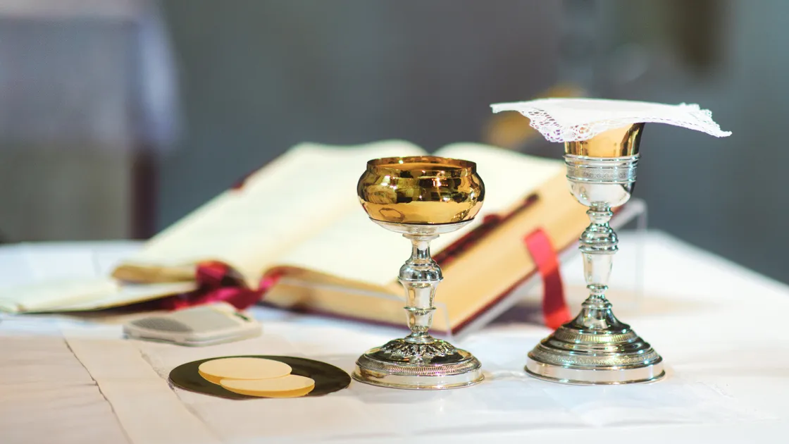 Cálice dourado, cibório de prata com véu de renda e duas hóstias sobre patena, ao lado de um missal aberto com fita vermelha, sobre toalha de altar branca.
