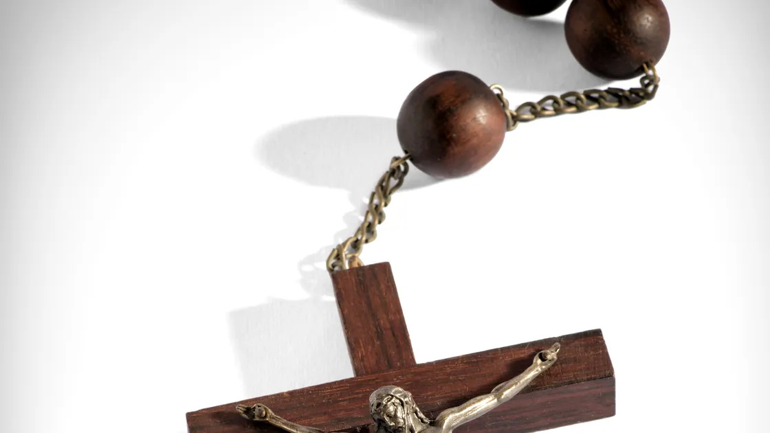 Terço de contas de madeira escura com crucifixo de madeira e corpo de Cristo em metal, em close-up sobre fundo branco.