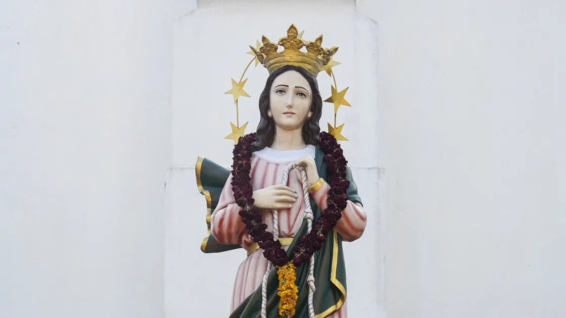 Estátua de Nossa Senhora Desatadora dos Nós com dois anjos segurando a corda de nós, muro branco ao fundo