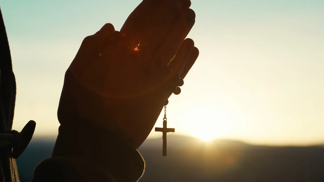 Mãos em oração segurando um terço com crucifixo, contraluz de pôr do sol ao fundo
