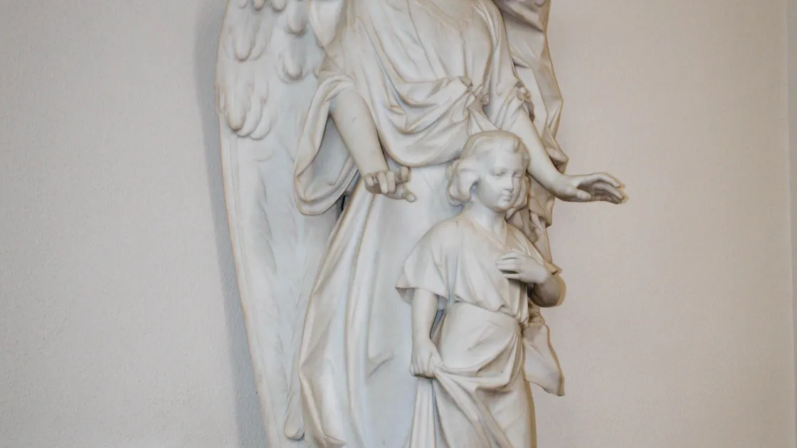 Escultura em mármore branco de um anjo da guarda amparando uma criança, sobre altar com velas votivas e flores