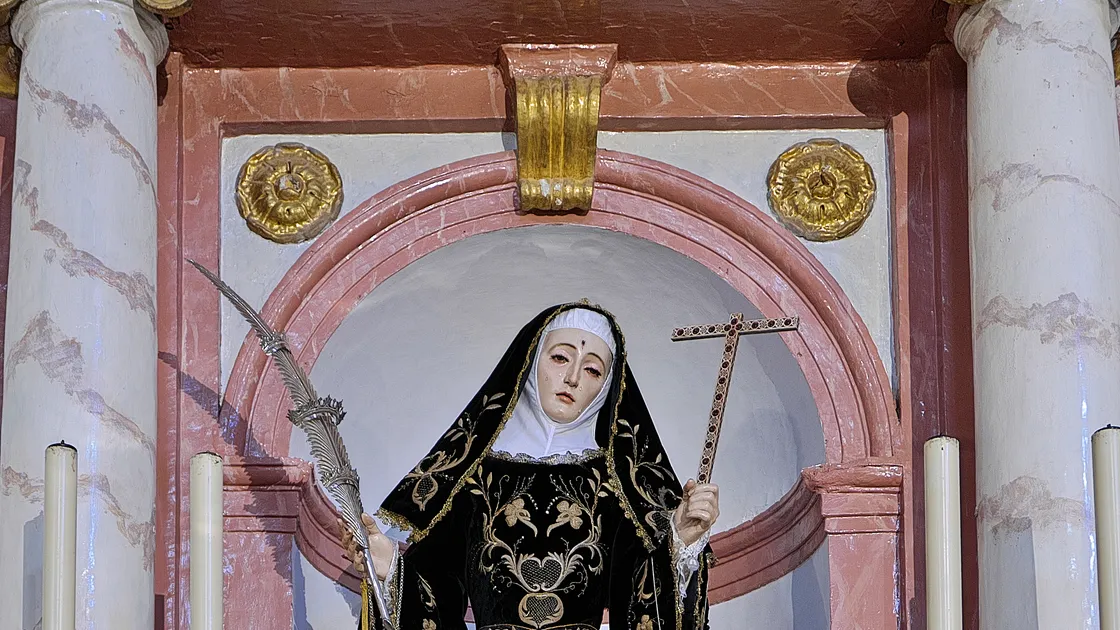 Escultura de Santa Rita de Cássia com hábito preto bordado a ouro, segurando cruz e ramo de palma, em altar de igreja