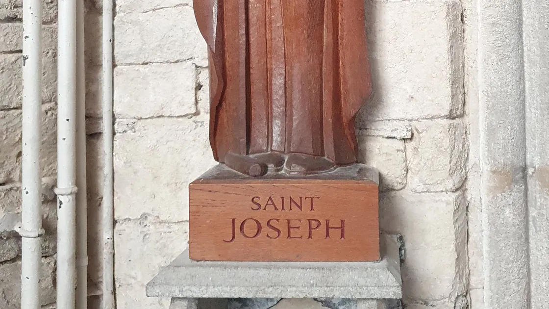 Escultura de madeira de São José com barba, mão direita erguida em bênção e a outra sobre o peito, sobre pedestal com a inscrição "Saint Joseph", junto a uma parede de pedra de igreja.