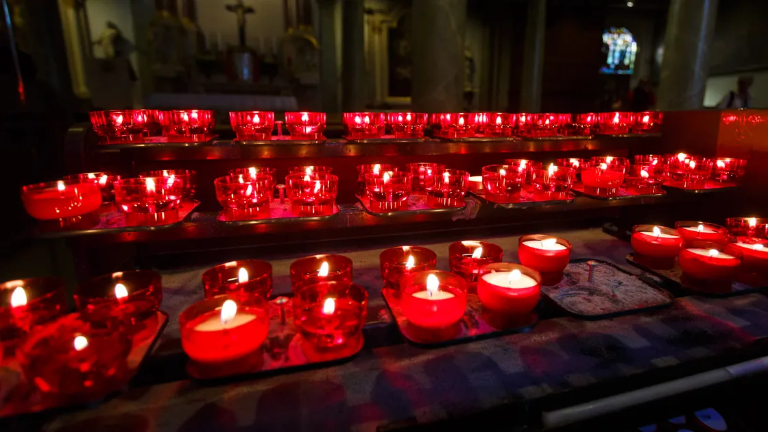 Fileiras de velas votivas vermelhas acesas numa capela católica, com crucifixo ao fundo do altar